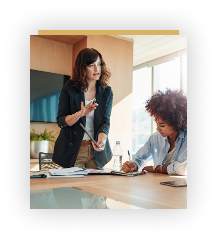 Two women in a modern office; one stands and speaks with a tablet in hand, while the other sits and takes notes at a conference table.